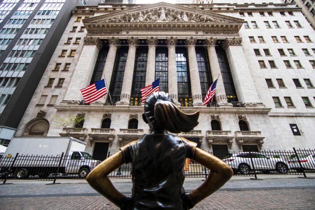 The Fearless Girl statue stands with hands on hips facing the New York Stock Exchange on Wall Street, American flags flying from the building's neoclassical facade