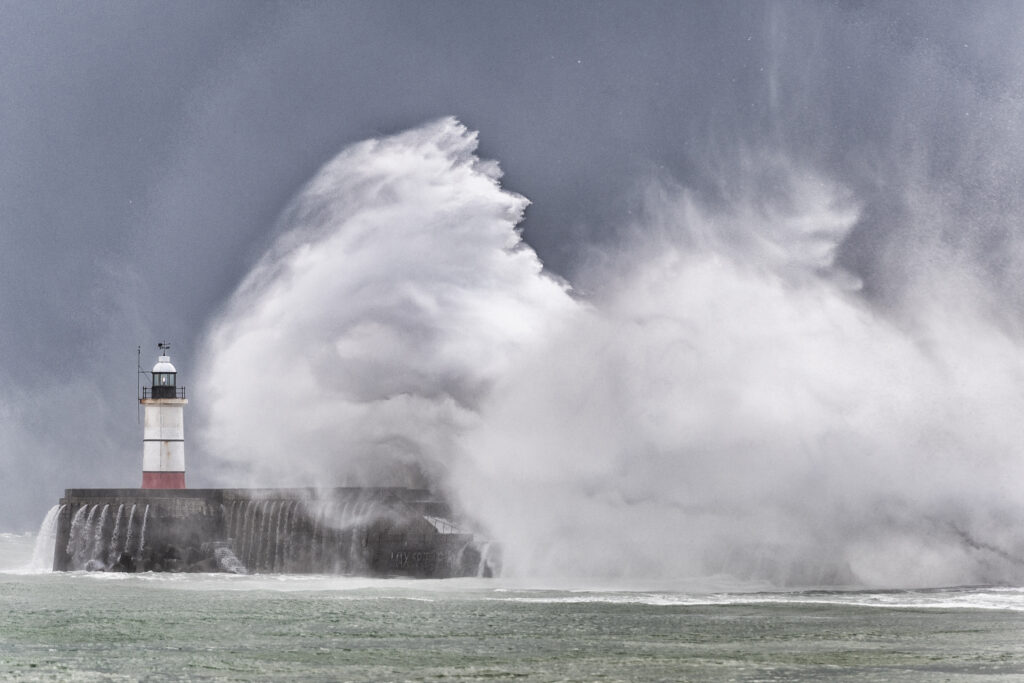 A massive wave crashes over a lighthouse and sea wall in a storm, dwarfing the structure against a grey sky.